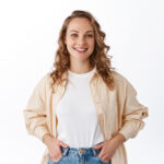 Home stylish young modern woman with blond hairdo, holding hands in jeans pockets and smiling carefree, looking confident and determined, white background