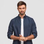 Home waist up portrait handsome serious unshaven male keeps hands together dressed dark blue shirt has talk with interlocutor stands against white wall self confident man freelancer